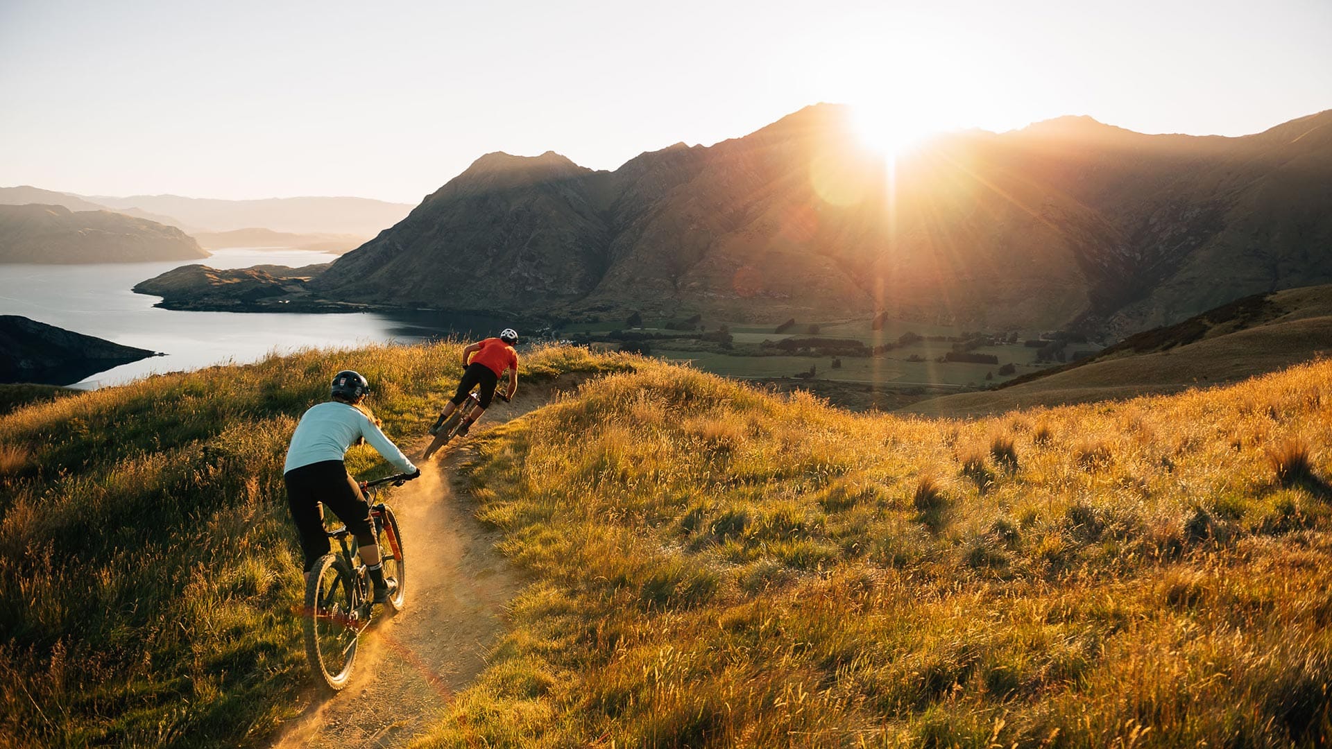 Two mountain bike riders on Bike Glendhu trail in golden hour overlooking Lake Wanaka and Roy's Peak, NZ